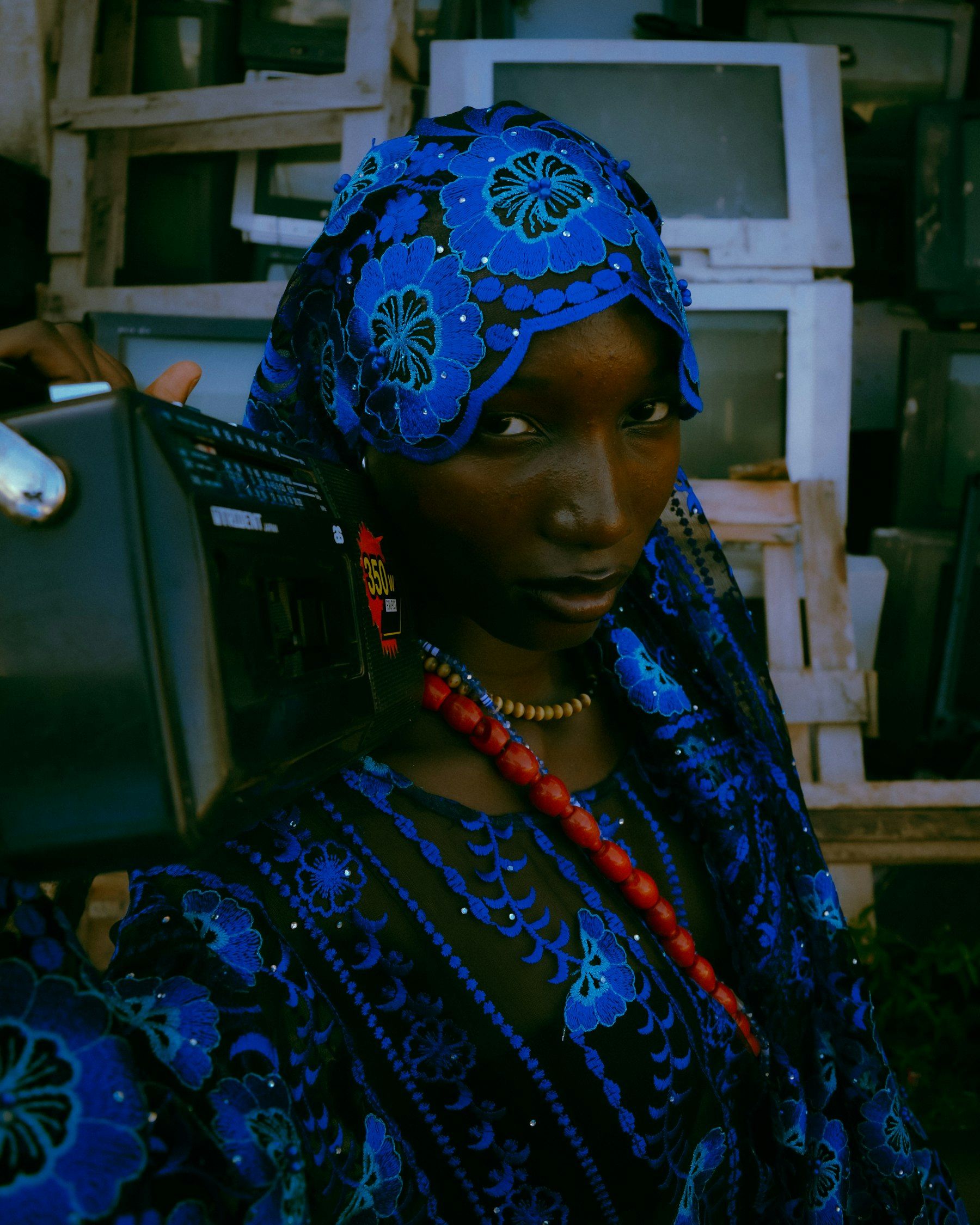 A woman in a blue patterned dress and headscarf, standing among old televisions.